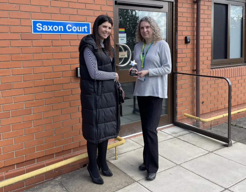 Two individuals standing outside a red brick building with a sign reading 'Saxon Court'. One person wears a long black vest over a striped shirt and black trousers, while the other wears a light grey jumper and black trousers, holding a star-shaped trophy